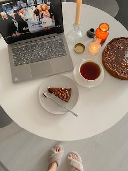 A cozy indoor scene featuring a slice of cake, cup of tea, and laptop on a round table.