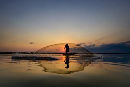 Free A serene silhouette of a fisherman casting a net on a calm lake at sunrise. Stock Photo