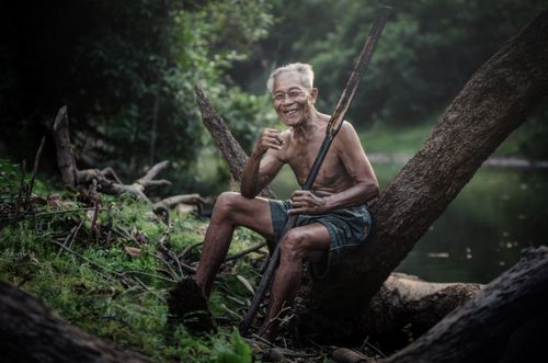 Free A senior man smiles happily while sitting on a log by a tranquil river, embodying peace. Stock Photo