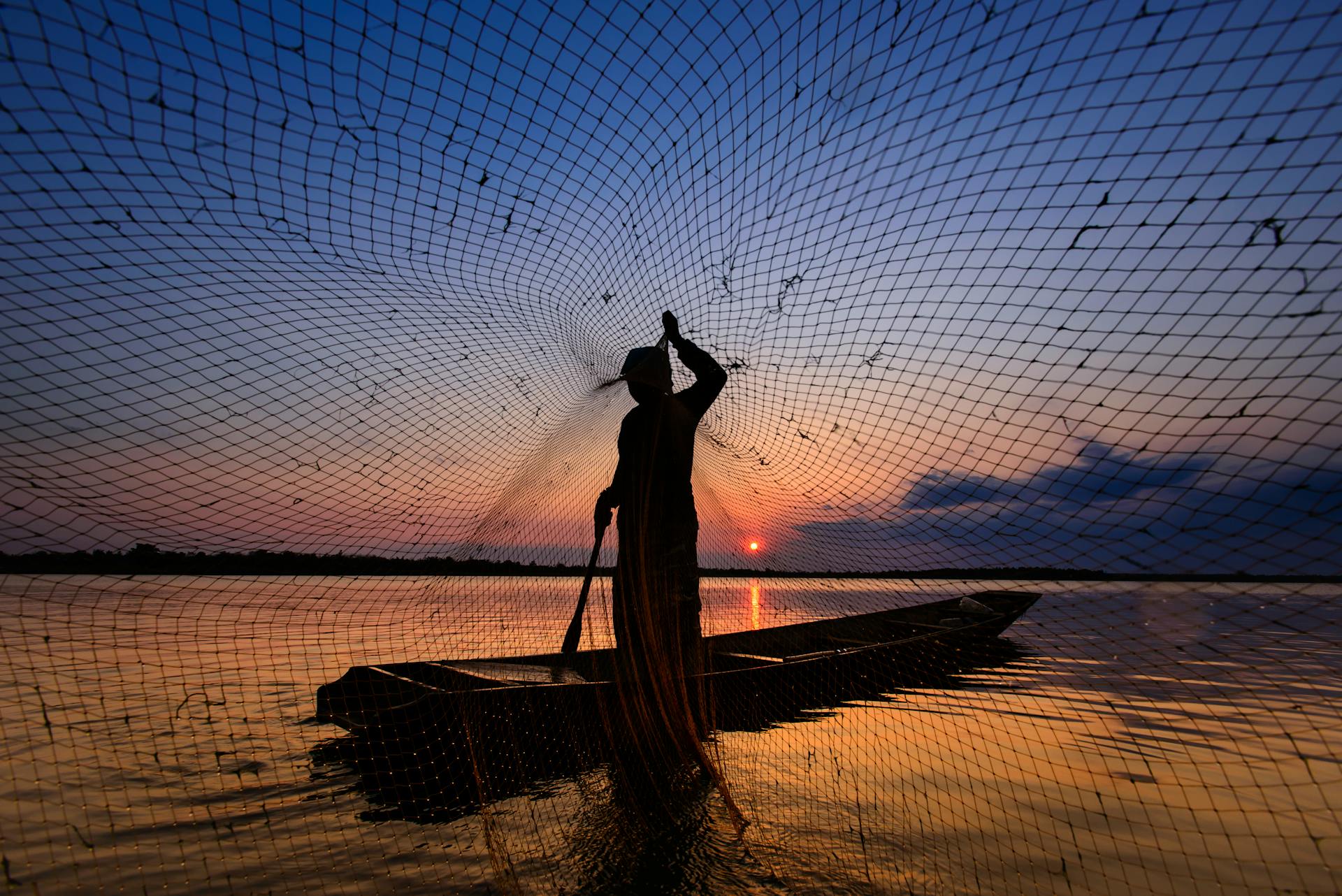 Silhouette of a fisherman casting a net from a boat on a tranquil body of water during a stunning sunset.