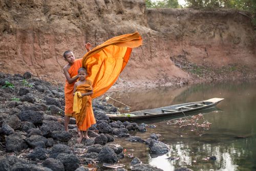 Free Two young monks in orange robes by a river, with a wooden boat and rocky shore. Stock Photo