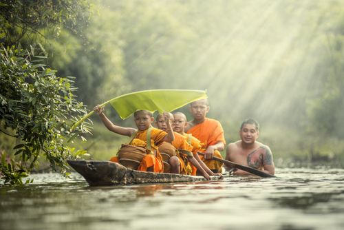 Free A group of young monks and a man paddling a wooden boat on a serene river, with sunrays filtering through trees. Stock Photo