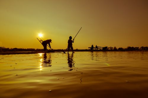 Free Silhouetted fishermen working at sunrise, casting shadows on calm waters reflecting the golden sky. Stock Photo