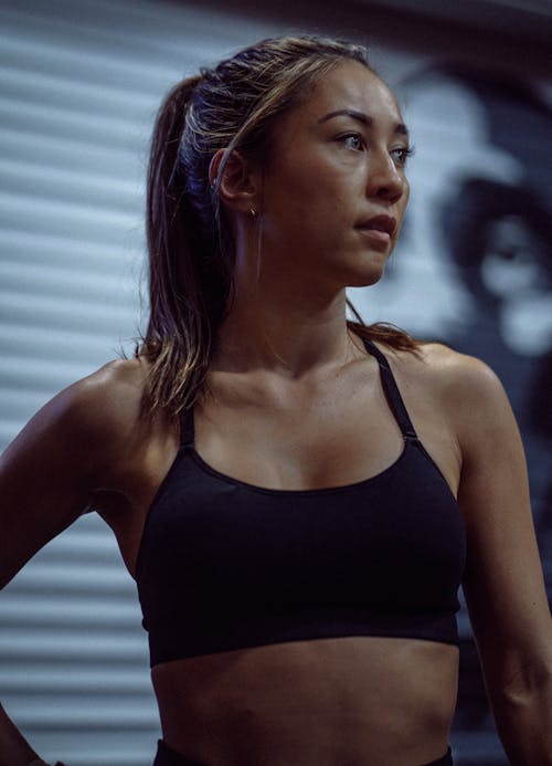 Serious sportswoman with ponytail in black top standing and looking away against white wall