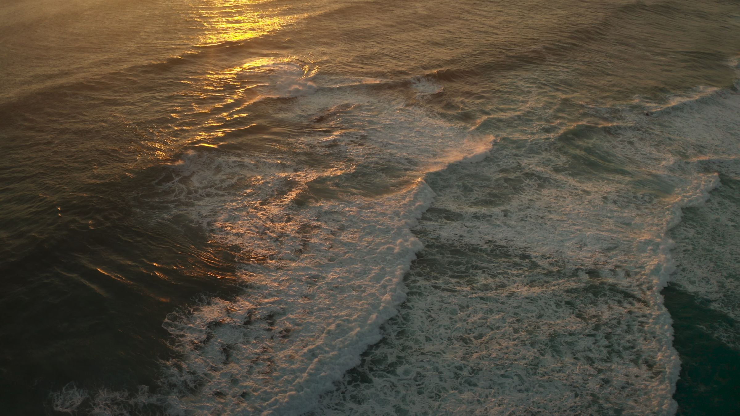 Aerial view of ocean waves at golden hour with the sun low over the horizon.