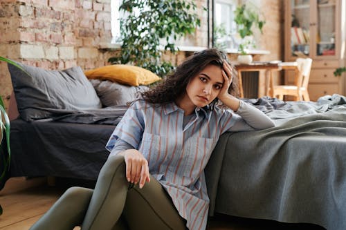 Free A Woman in Striped Shirt Sitting Near the Bed with Her Hand on Her Head Stock Photo