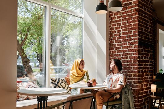 A bearded man and woman in hijab conversing in a sunlit café by a glass window.