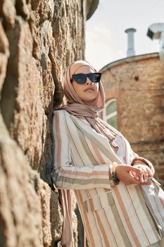 Fashionable woman in a striped jacket and hijab, leaning against a rustic stone wall outdoors.
