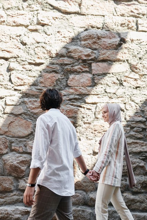 Free A couple walks hand in hand along a rustic stone wall, exuding togetherness in an outdoor setting. Stock Photo