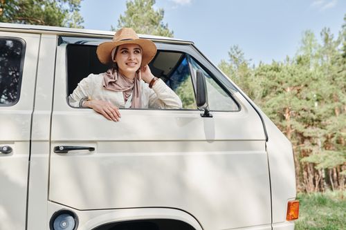 Free Smiling woman in sun hat leaning out of van window amidst a forest setting. Stock Photo