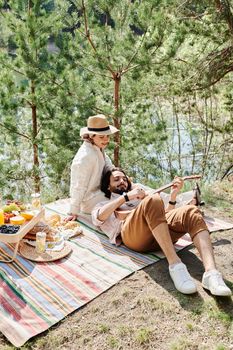 Couple enjoying a sunny picnic by a lake with guitar music in a serene outdoor setting.