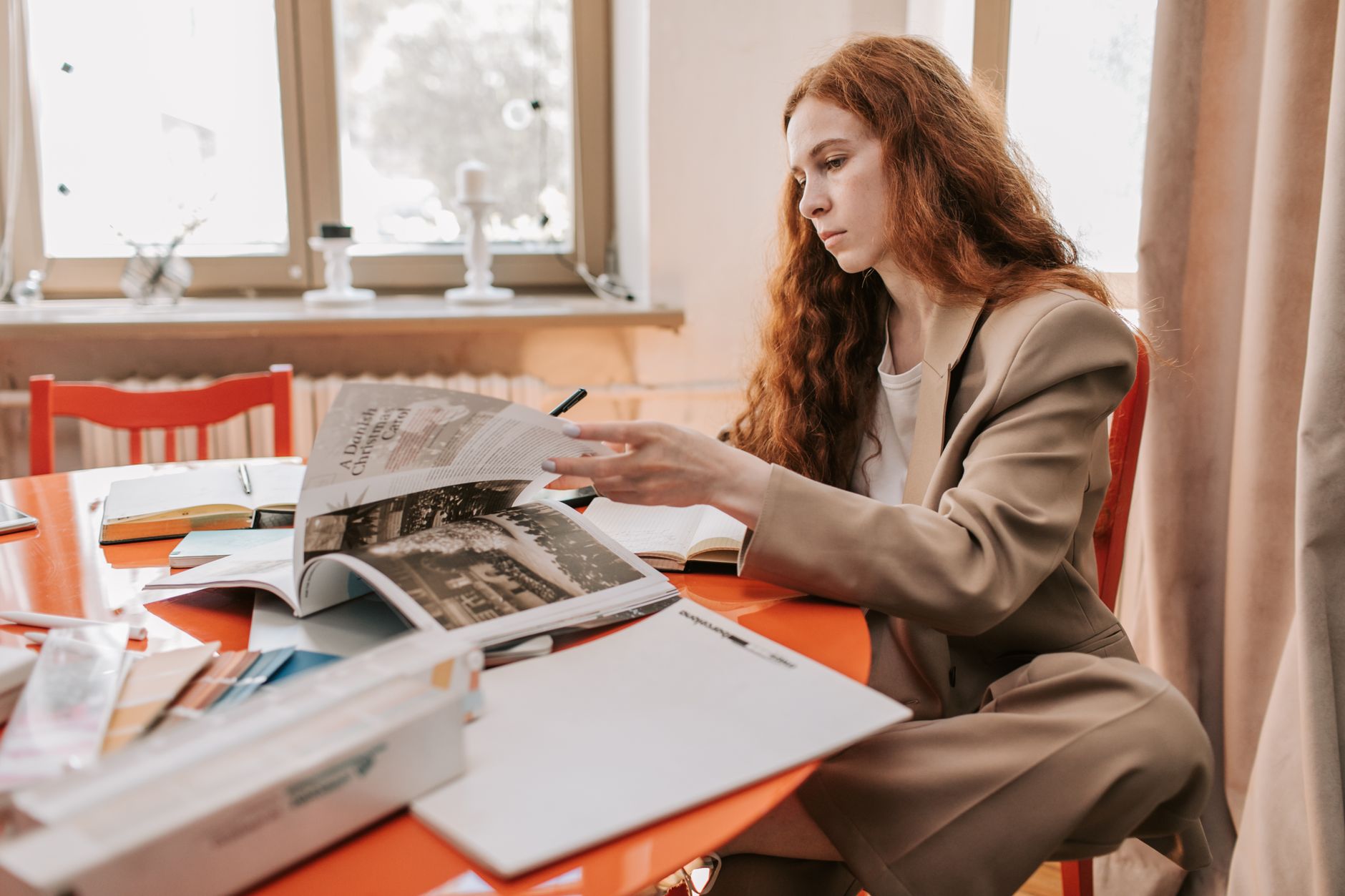 A focused woman in a beige suit reading documents at a desk in a modern office setting.