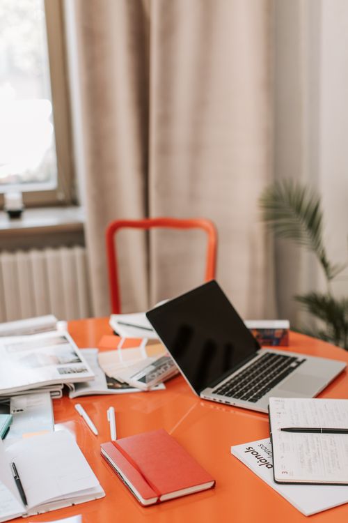 Free A modern office desk setup with a laptop, notebooks, and stationery on a bright orange table. Stock Photo