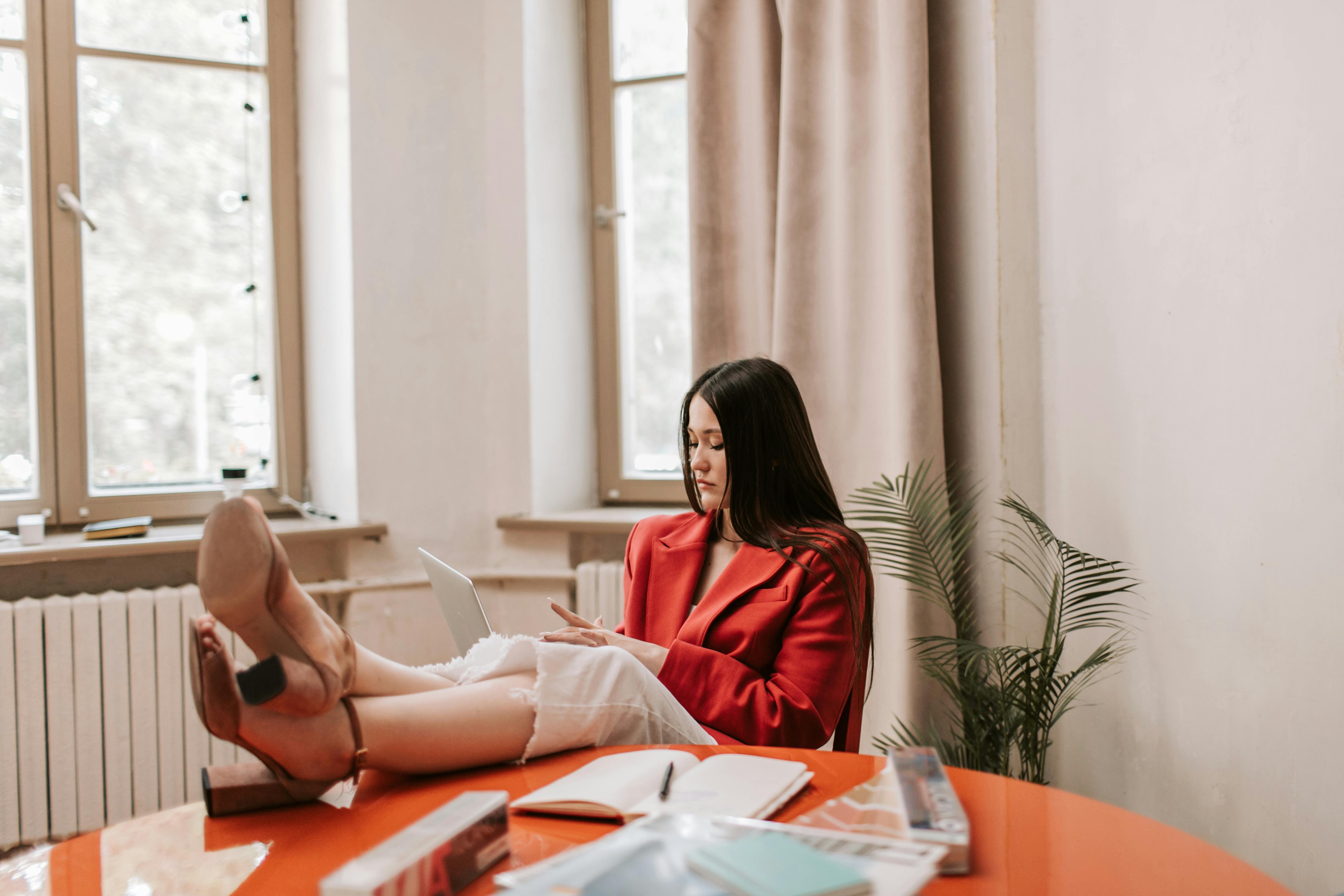 A woman in a red blazer relaxes with her feet on the office table, working on a laptop.
