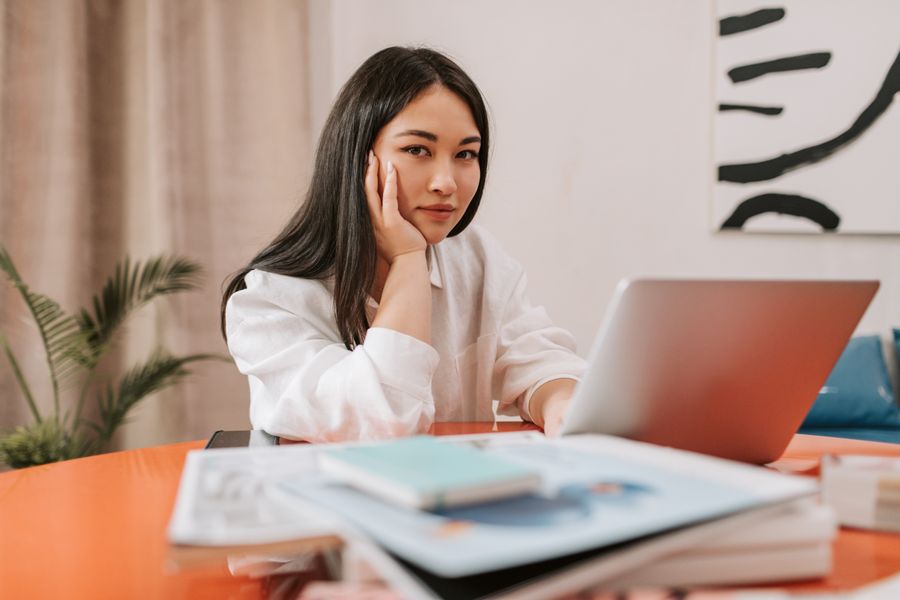 Young Asian woman sitting at desk with her hand on cheek, in front of a laptop, surrounded by books.
