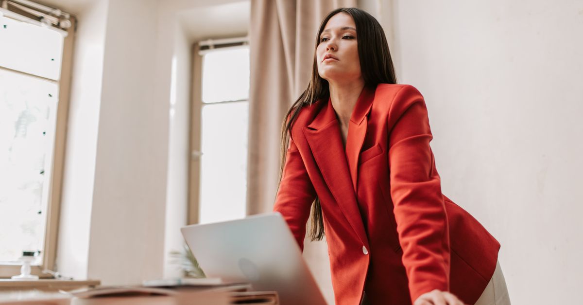 Businesswoman in a red blazer leaning over a desk with a laptop, exuding confidence.