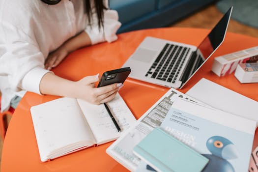 Woman multitasking with a smartphone and laptop at an office desk, engaging in digital work.