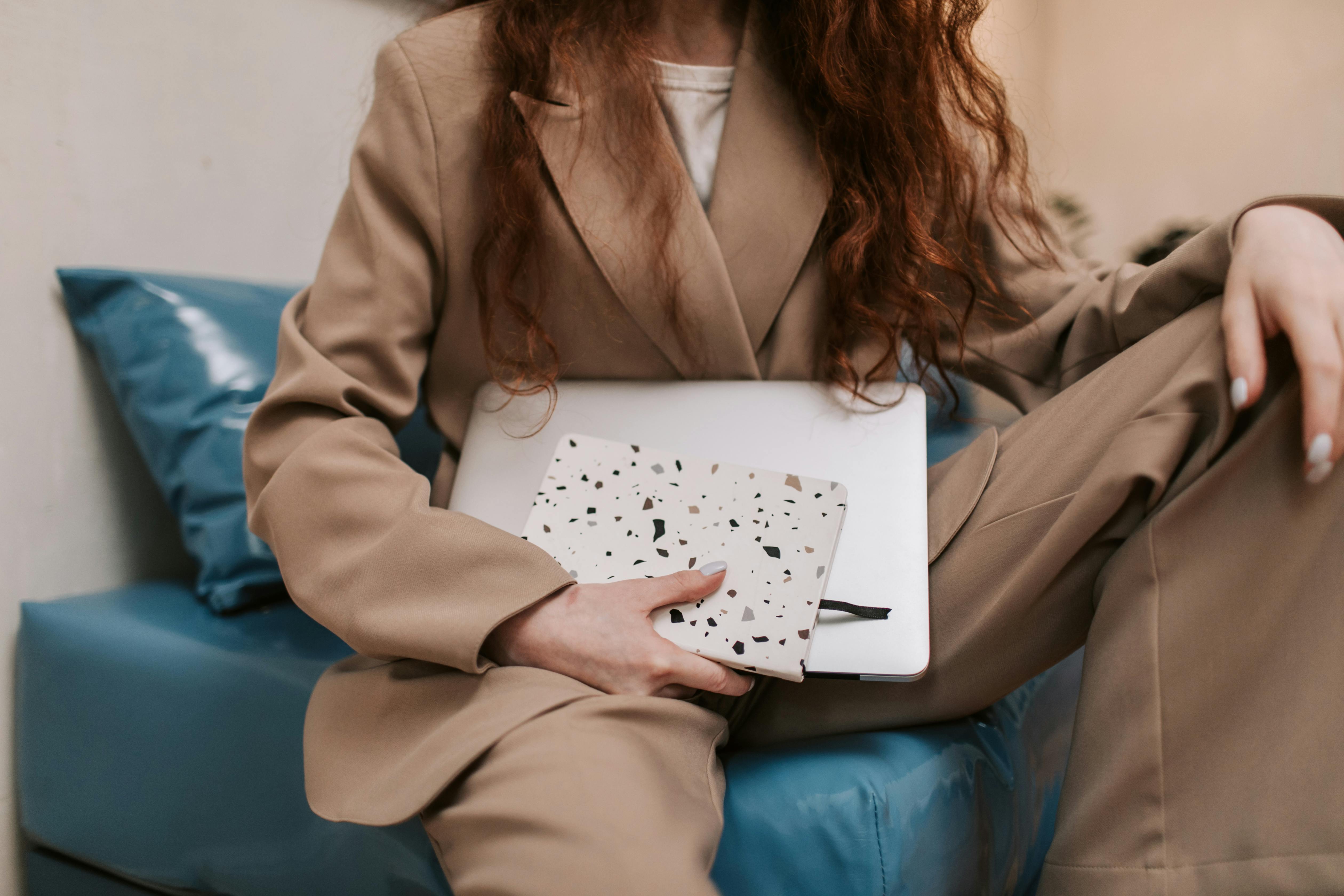 Businesswoman sitting with a laptop on a sofa, holding a notebook indoors.