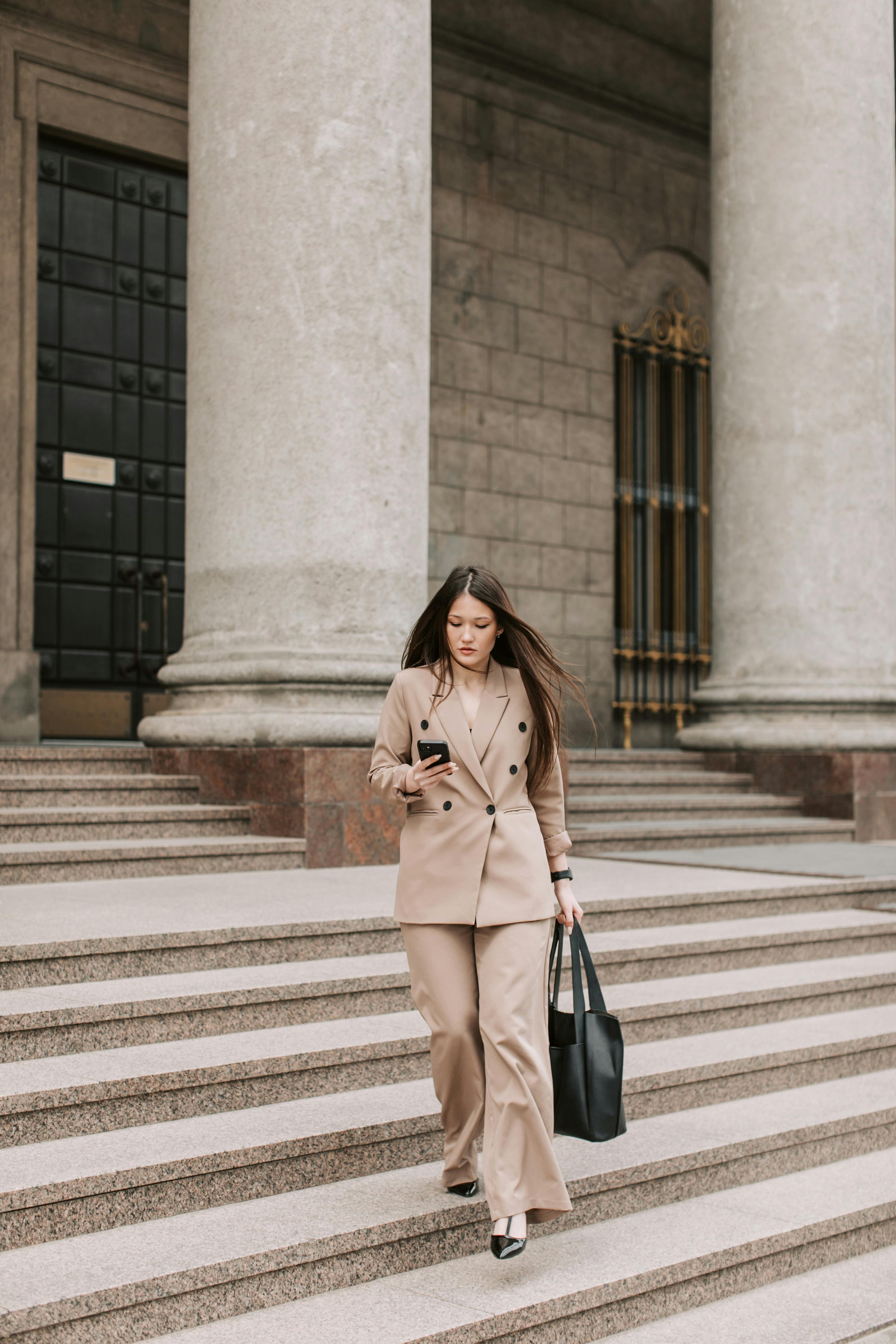 woman in beige blazer carrying bag walking down the concrete stairs while using her cellphone