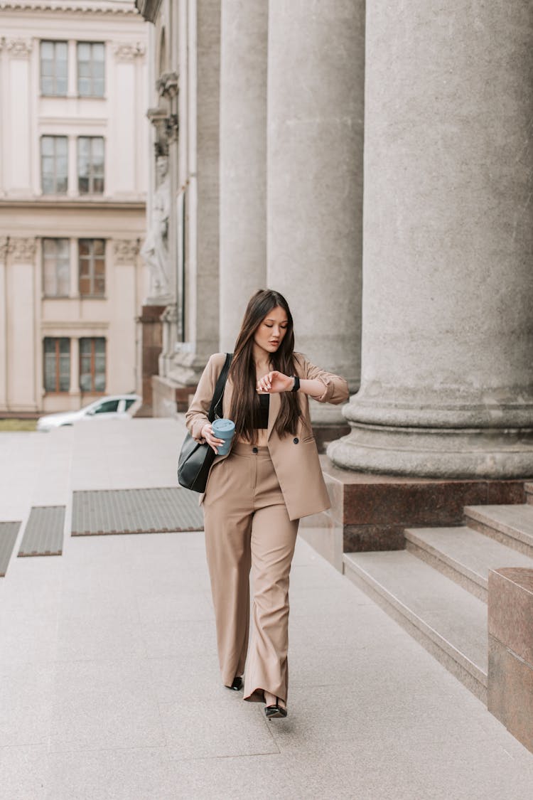 Woman In Beige Corporate Attire Looking At Her Wristwatch While Walking Near The Pillars Of A Building