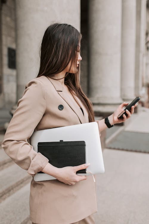 A Woman Using Her Phone while Carrying a Laptop and a Notebook