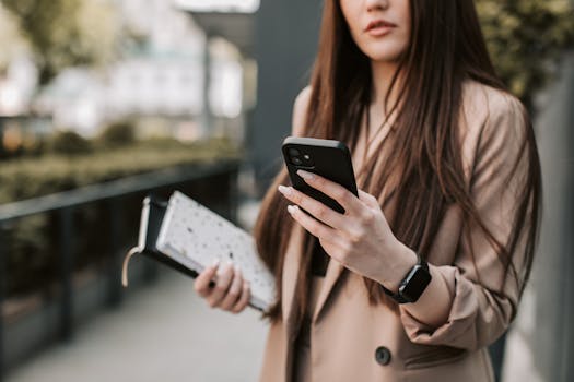 Young woman in beige blazer checking smartphone while holding a notebook outside.