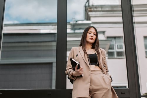 Free Stylish Asian woman in sharp corporate attire posing confidently against a glass wall. Stock Photo