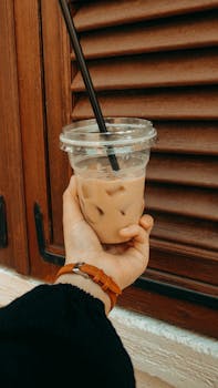 Hand holding iced coffee with straw outdoors beside a wooden window shutter, a perfect refreshing drink.