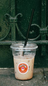 A refreshing iced coffee in a disposable cup with a straw against a textured green gate background.