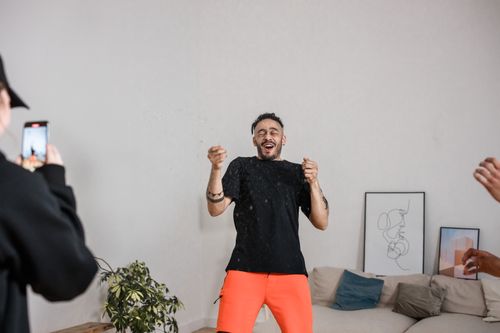 Free A man in bright shorts laughs cheerfully as friends film him inside a cozy room. Stock Photo