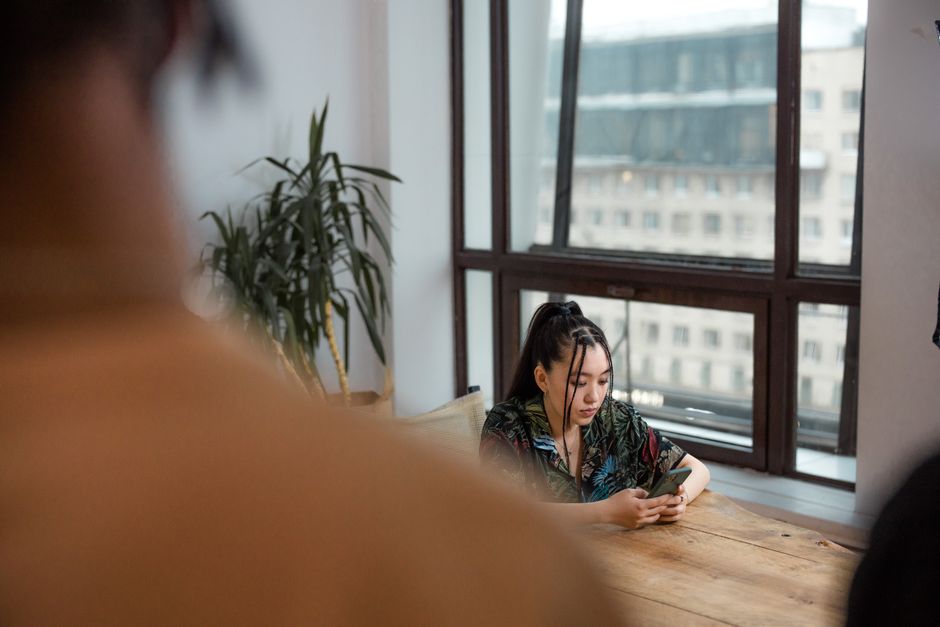 Young woman engrossed with a smartphone while sitting indoors by a large window.