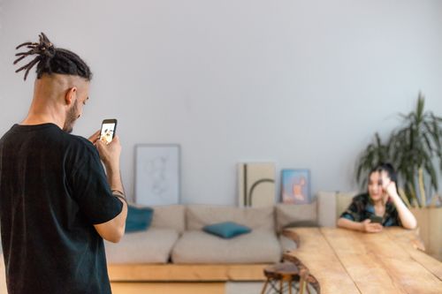 Free A man using a smartphone to capture a woman sitting in a modern living room. Stock Photo