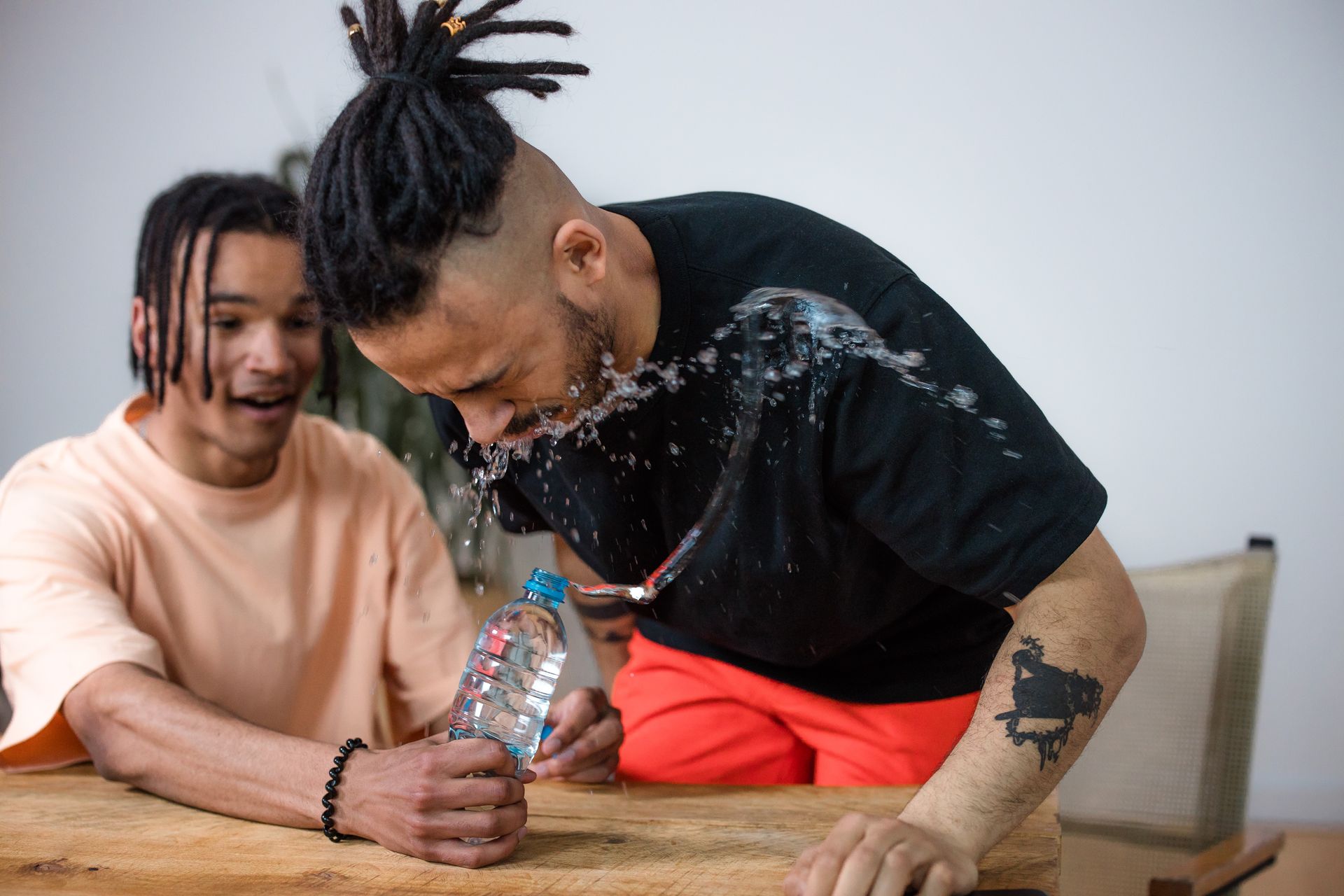 Two adult friends laughing during a playful water splash prank at a wooden table indoors.
