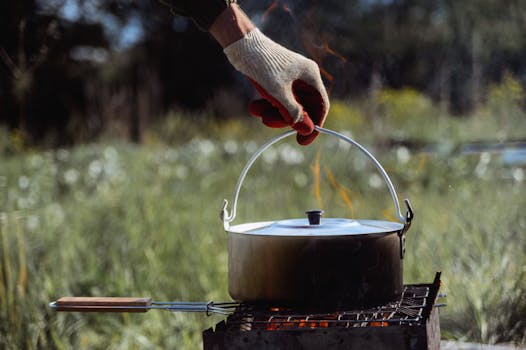 A close-up of a person in gloves handling a pot on a campfire grill outdoors.