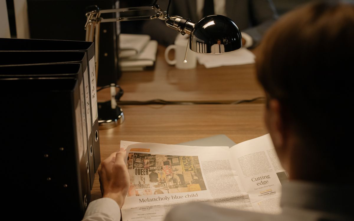 A person reads a newspaper at a desk with a lamp, suggesting investigation work.