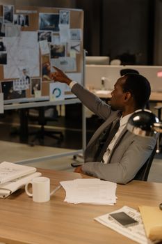 An African American man in a suit points at a board full of evidence in an office setting.