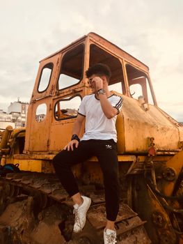 Stylish young man in white shirt sitting on a vintage yellow excavator outdoors.