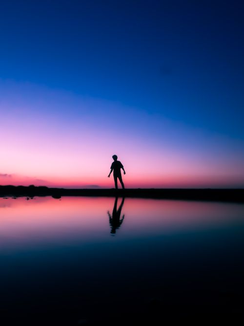Free A serene silhouette of a person standing by water during a colorful twilight sky. Stock Photo