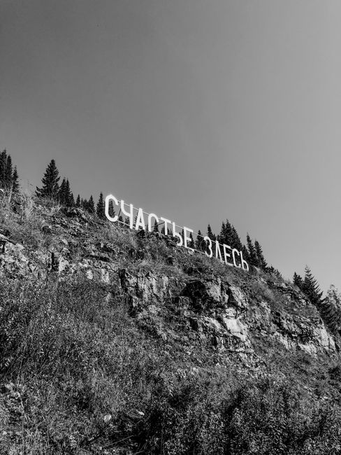 Black and white photo of a hillside with Cyrillic sign reading 'happiness here'.