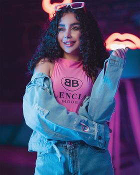 Fashionable woman with curly hair poses in denim jacket against neon backdrop.