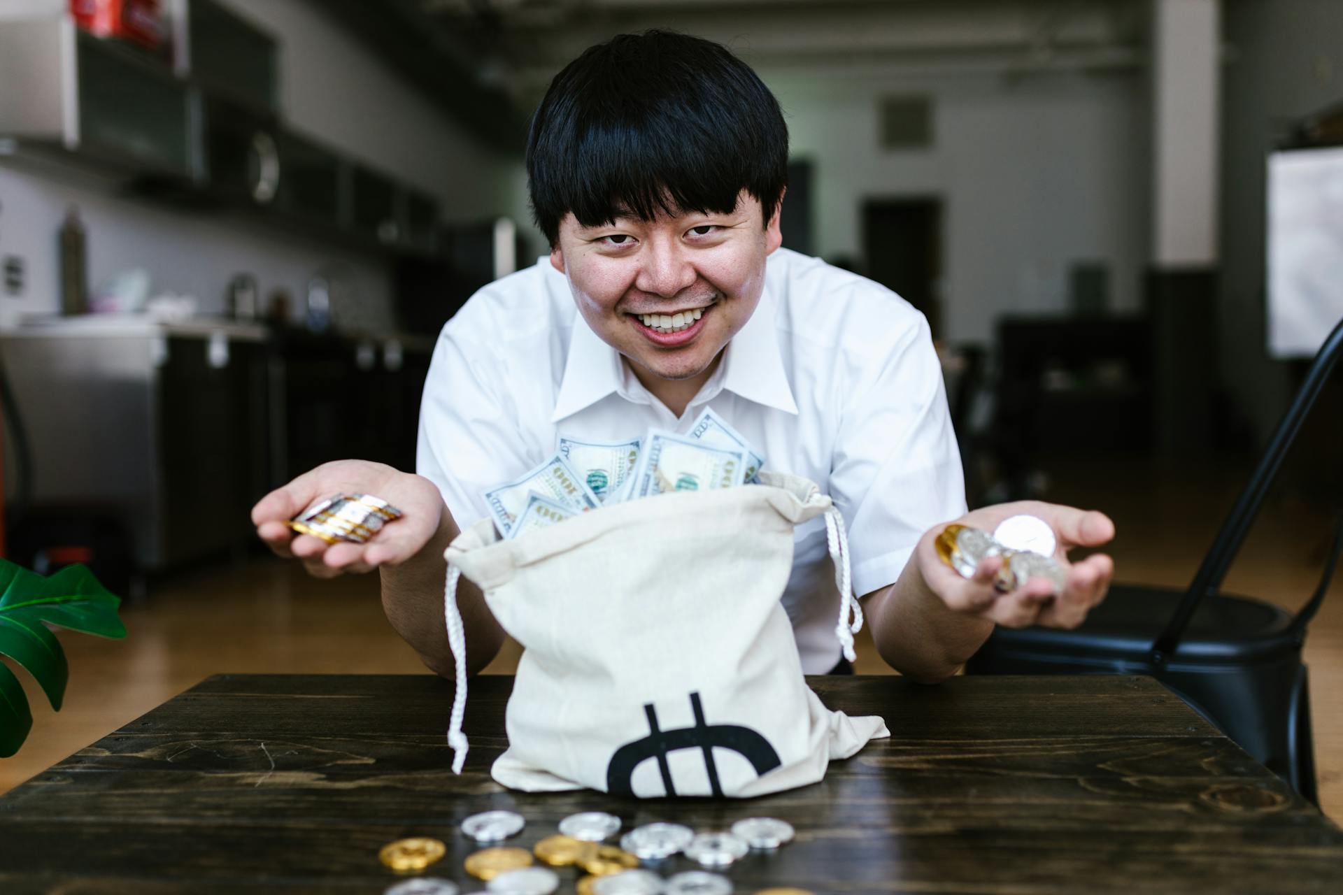 Asian man smiling with cryptocurrency coins and cash at home office setting.