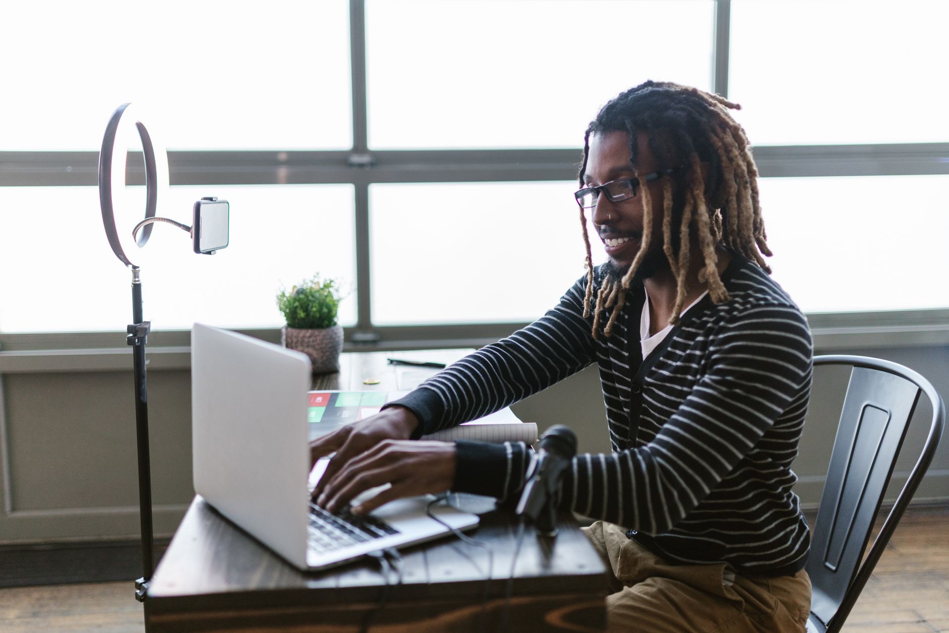 African American man with dreadlocks working on laptop indoors, using ring light and microphone.
