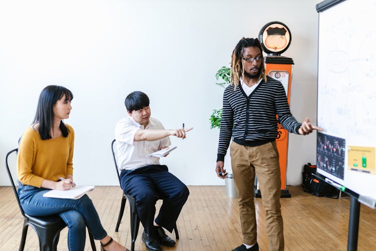 A Man In Black And White Stripes Teaching A Man And A Woman Sitting On A Chair