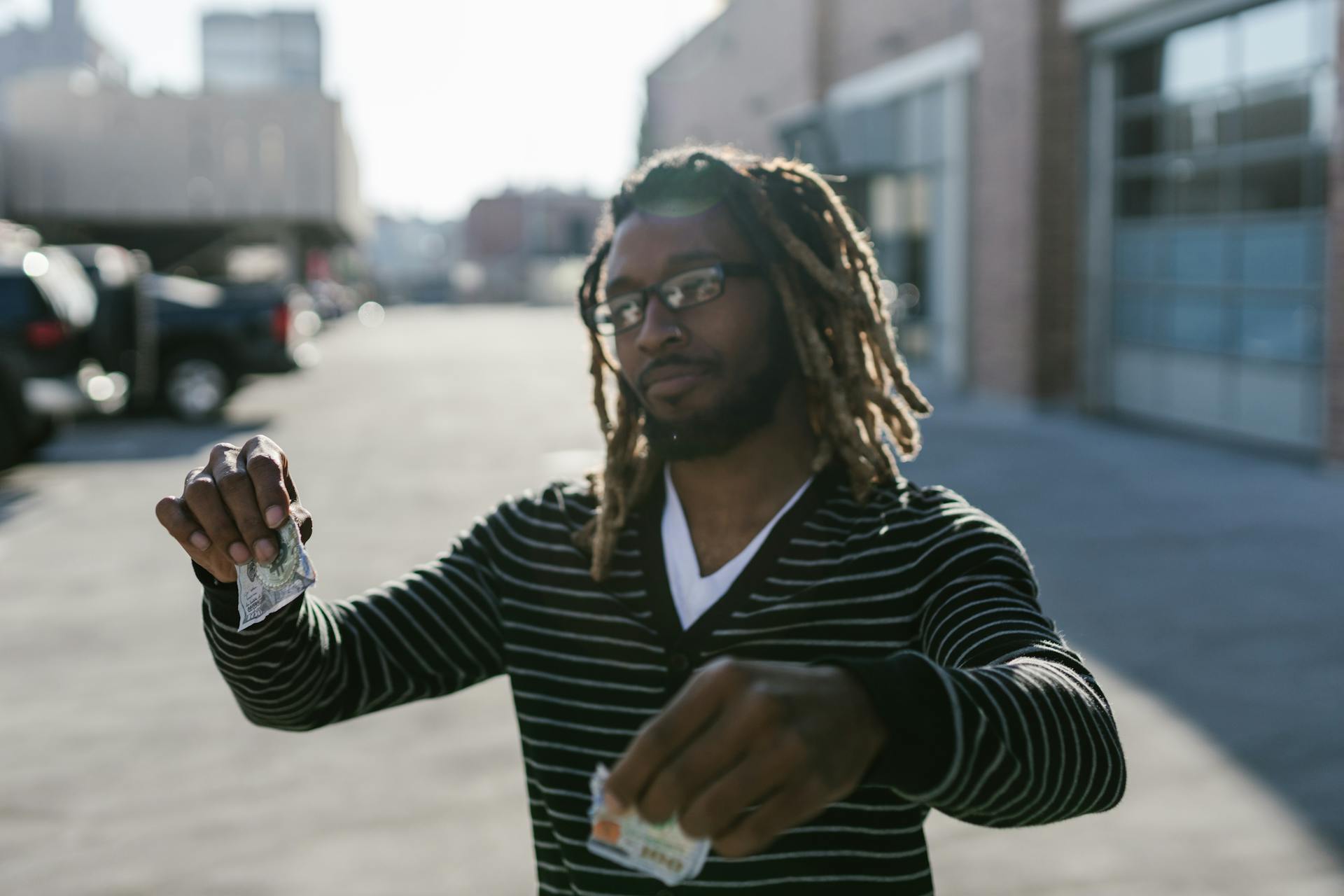 A young man with glasses and dreadlocks tearing a banknote in an urban outdoor setting.