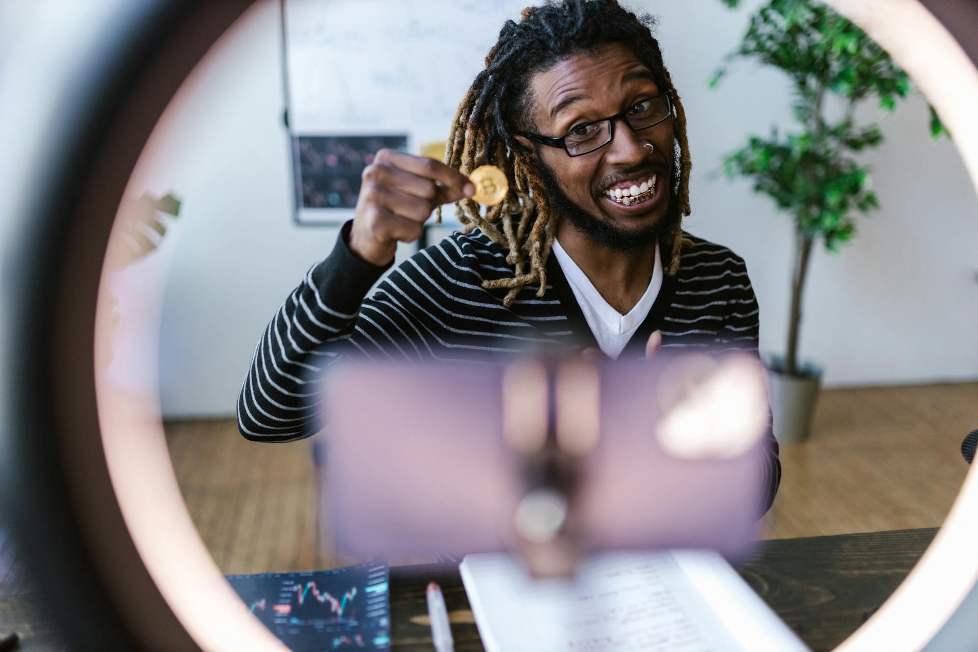 African American man smiling while holding a Bitcoin in front of a smartphone for a video recording.