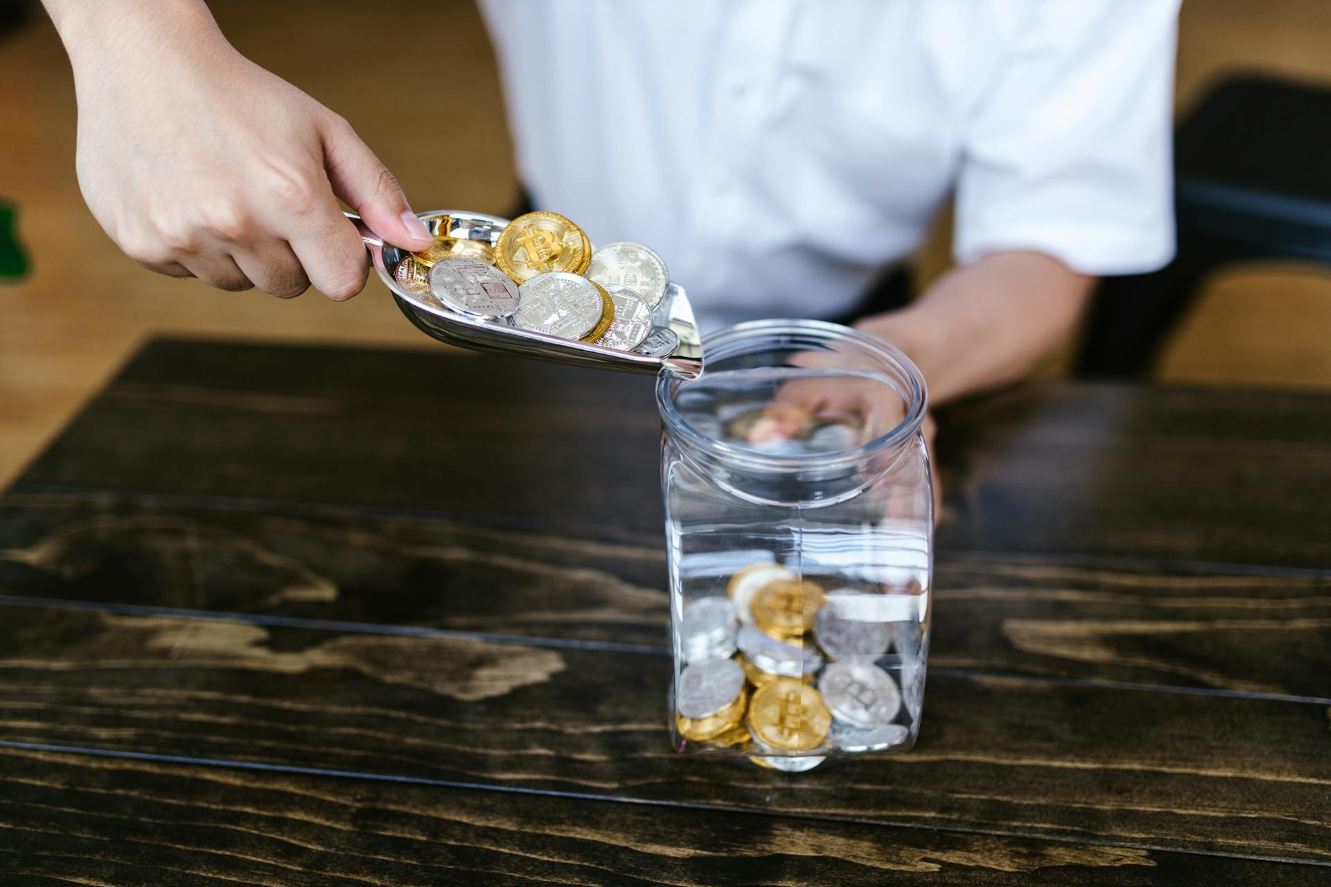 Hand placing gold and silver coins into a glass jar on a wooden table indoors.