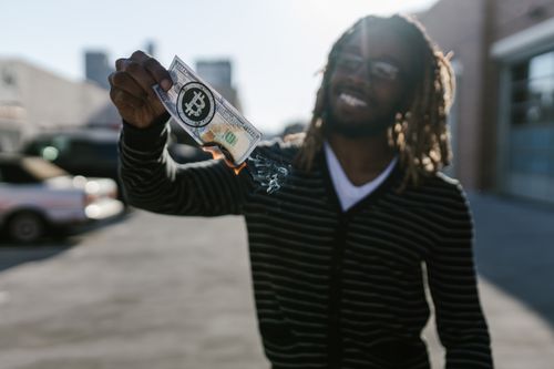 Free A man with dreadlocks holds a burning bill featuring a bitcoin logo, symbolizing cryptocurrency volatility. Stock Photo