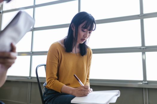 A woman in a yellow sweater writes in a notebook inside a bright room.