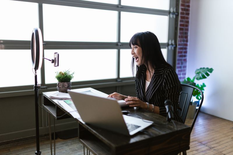 Asian woman in office setting recording content with laptop and ring light, showcasing elegant business setup.