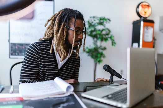 A young man with dreadlocks and eyeglasses records a podcast at a desk with a microphone and laptop.
